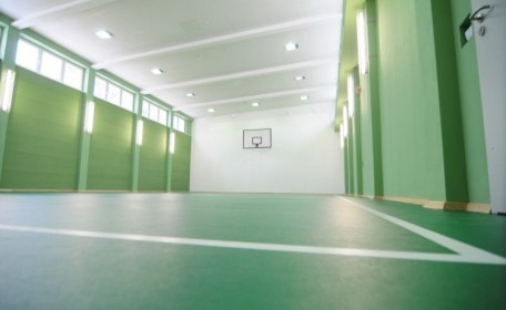 Zielona podłoga hali sportowej z białymi liniami, widok na kosz do koszykówki na białej ścianie w tle, oświetlenie jarzeniowe