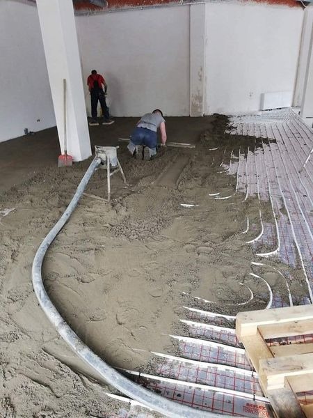 Aplikacja wylewki jastrychowej na ogrzewanie podłogowe z widoczną pompą, dwoma pracownikami i częściowo wyrównaną powierzchnią w surowym wnętrzu.