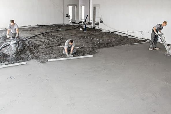 Trzech pracowników wyrównuje i wygładza wylewkę podłogową w dużym pomieszczeniu; jeden używa poziomicy, a dwaj pozostali zacierają powierzchnię mechanicznymi zacieraczkami.