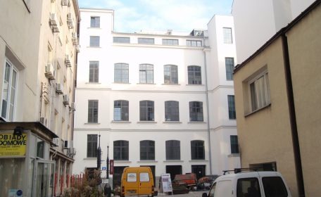 Biały budynek wielopiętrowy z ciemnymi oknami w otoczeniu innych budynków, widoczny samochód dostawczy i materiały budowlane na placu