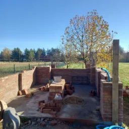 Wznoszenie murów z czerwonej cegły na betonowej wylewce, widoczne zbrojenia i niebieski przewód wentylacyjny, w tle jesienne drzewo i zabudowa jednorodzinna.