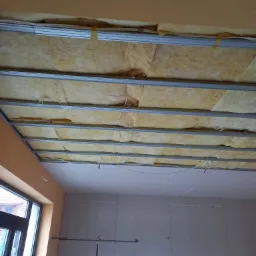 Montaż sufitu podwieszanego: widok na metalową konstrukcję z izolacją z wełny mineralnej, częściowo zakryte płytami gipsowo-kartonowymi, widoczne przewody elektryczne.