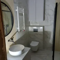 Nowoczesna łazienka z marmurowymi płytkami, okrągłym lustrem i minimalistyczną umywalką nablatową, widoczny grzejnik drabinkowy i kabina prysznicowa z czarnymi profilami.