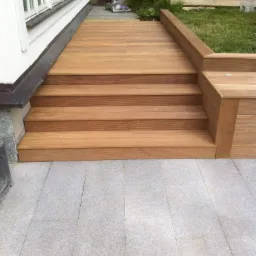 Nowoczesny taras z drewnianymi schodami i siedziskiem, wykończony szarą kostką granitową, przylegający do białego budynku z oknami.