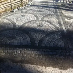 Ułożona kostka granitowa w łuki, z widocznymi śladami po deszczu i drewnianym płotem w tle.