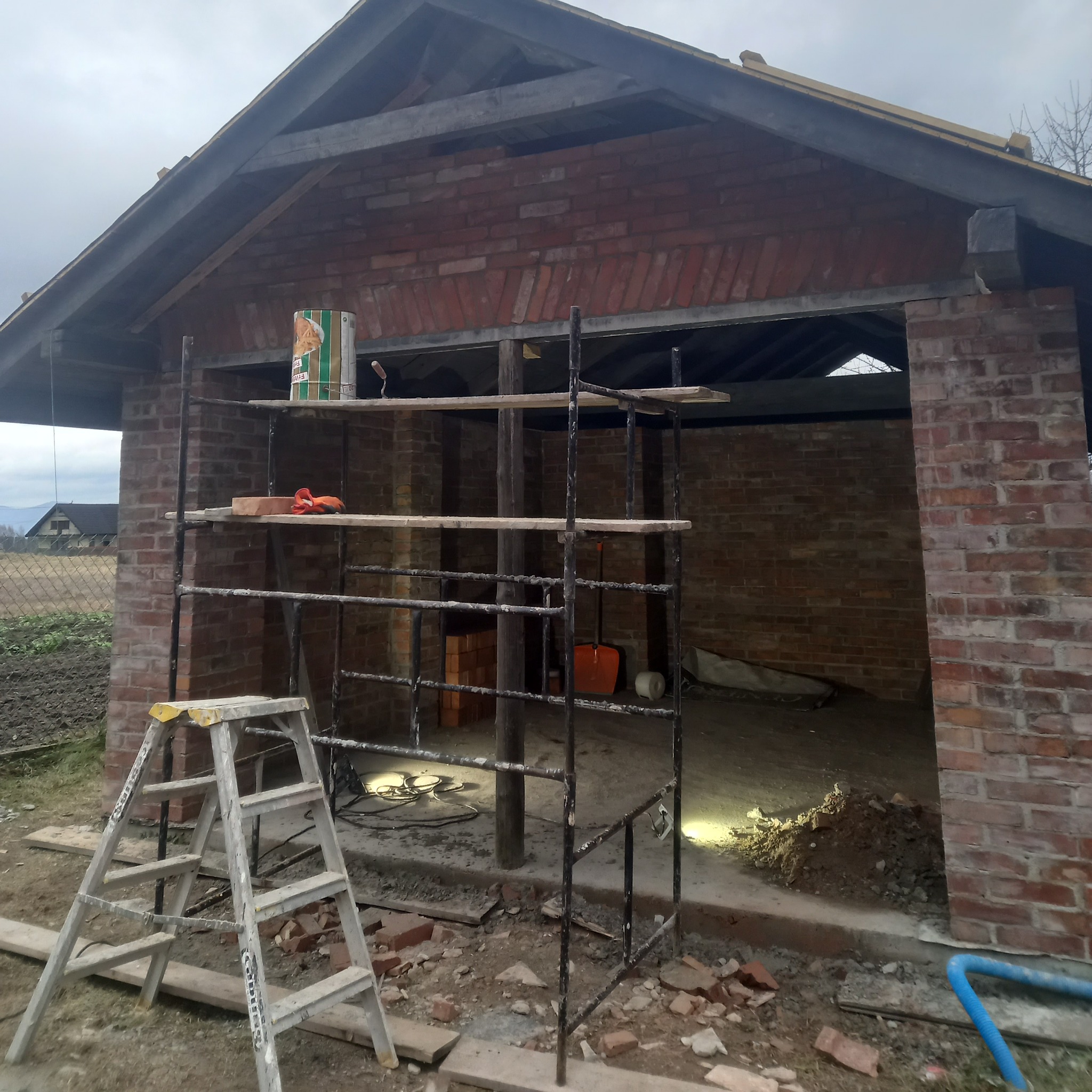 Budowa murowanej konstrukcji z czerwonej cegły, widoczne rusztowanie, drabina aluminiowa, wiadro z zaprawą murarską oraz porozrzucane fragmenty cegieł na ziemi.