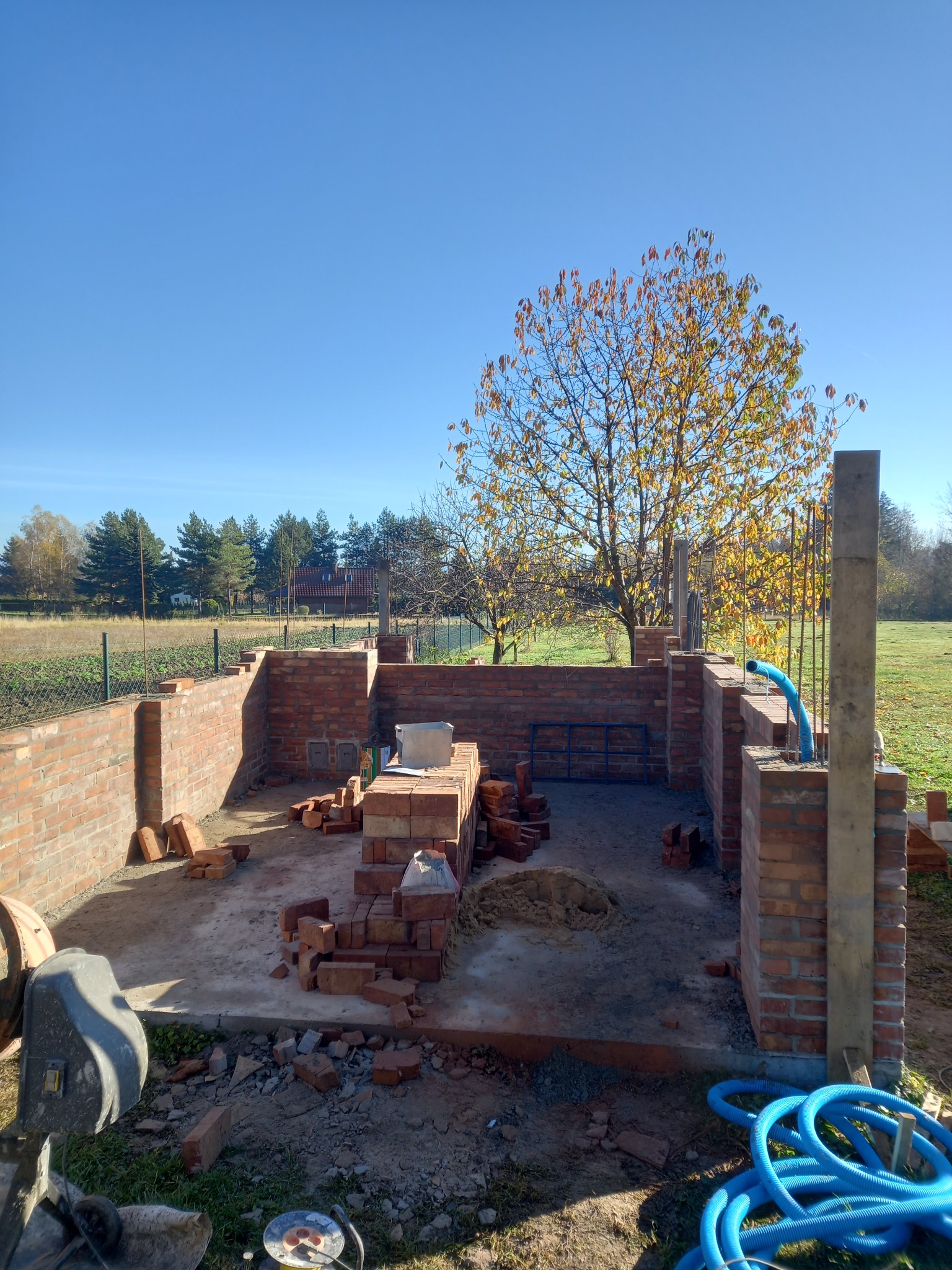 Wznoszenie murów z czerwonej cegły na betonowej wylewce, widoczne zbrojenia i niebieski przewód wentylacyjny, w tle jesienne drzewo i zabudowa jednorodzinna.