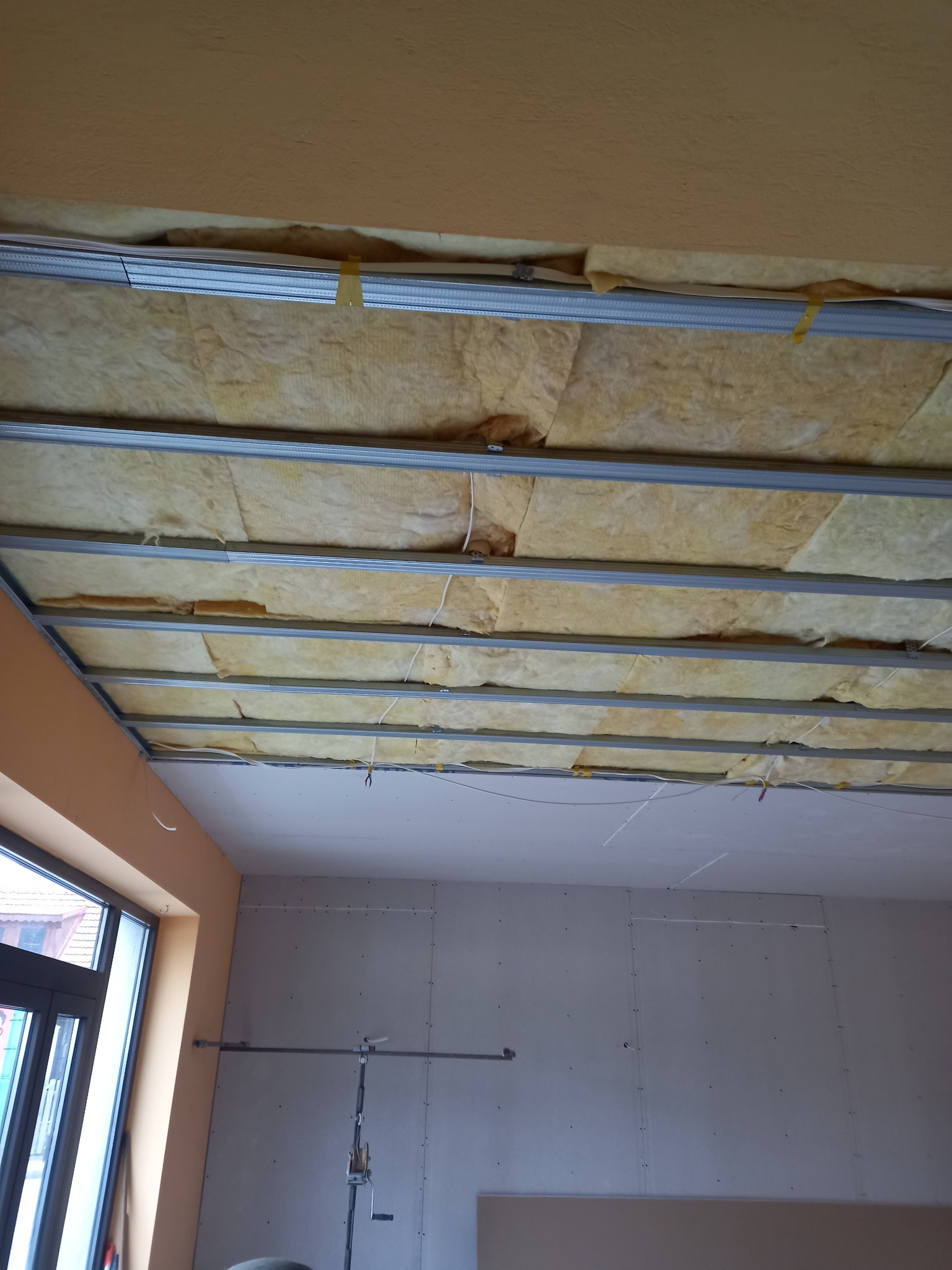 Montaż sufitu podwieszanego: widok na metalową konstrukcję z izolacją z wełny mineralnej, częściowo zakryte płytami gipsowo-kartonowymi, widoczne przewody elektryczne.