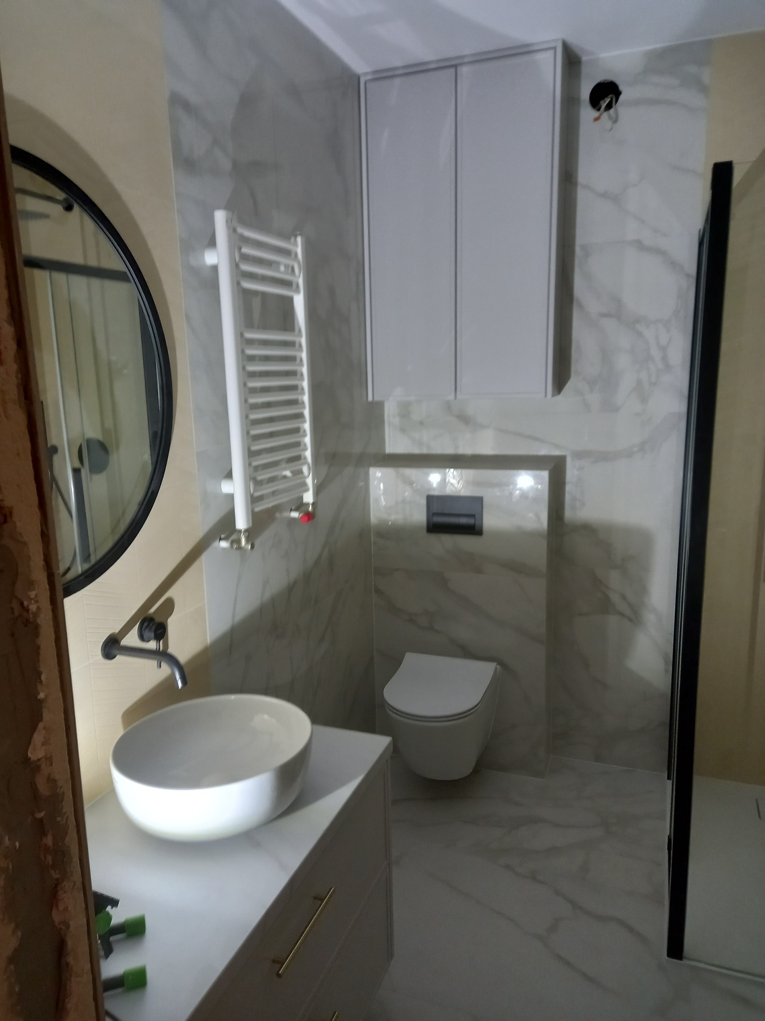 Nowoczesna łazienka z marmurowymi płytkami, okrągłym lustrem i minimalistyczną umywalką nablatową, widoczny grzejnik drabinkowy i kabina prysznicowa z czarnymi profilami.