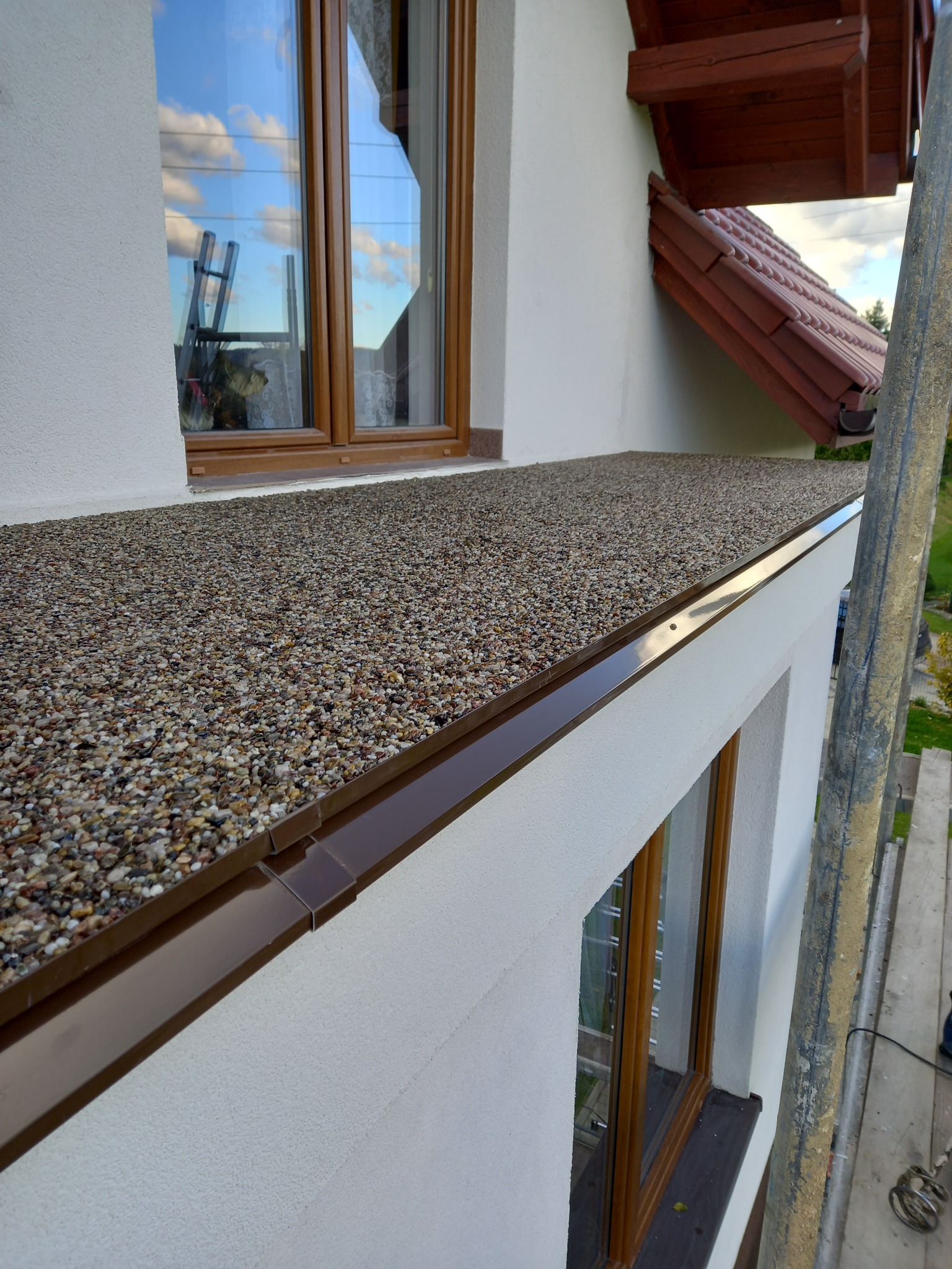 Wykończony taras z żywicy kamiennej na zewnątrz budynku, widoczna faktura drobnych kamyczków, brązowe obramowanie, okno z odbiciem drabiny i fragment dachu z dachówką.