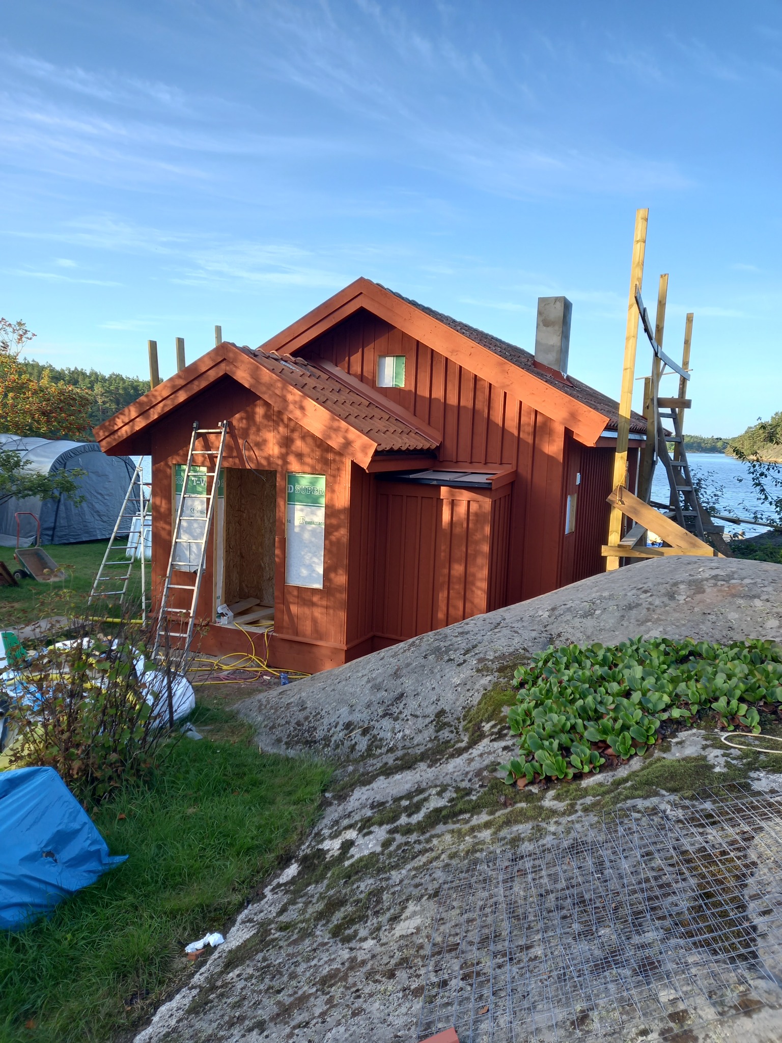 Czerwony, drewniany domek letniskowy w trakcie budowy w Jeleśni, z widocznymi rusztowaniami i drabiną opartą o wejście, położony na skalistym terenie z widokiem na jezioro.