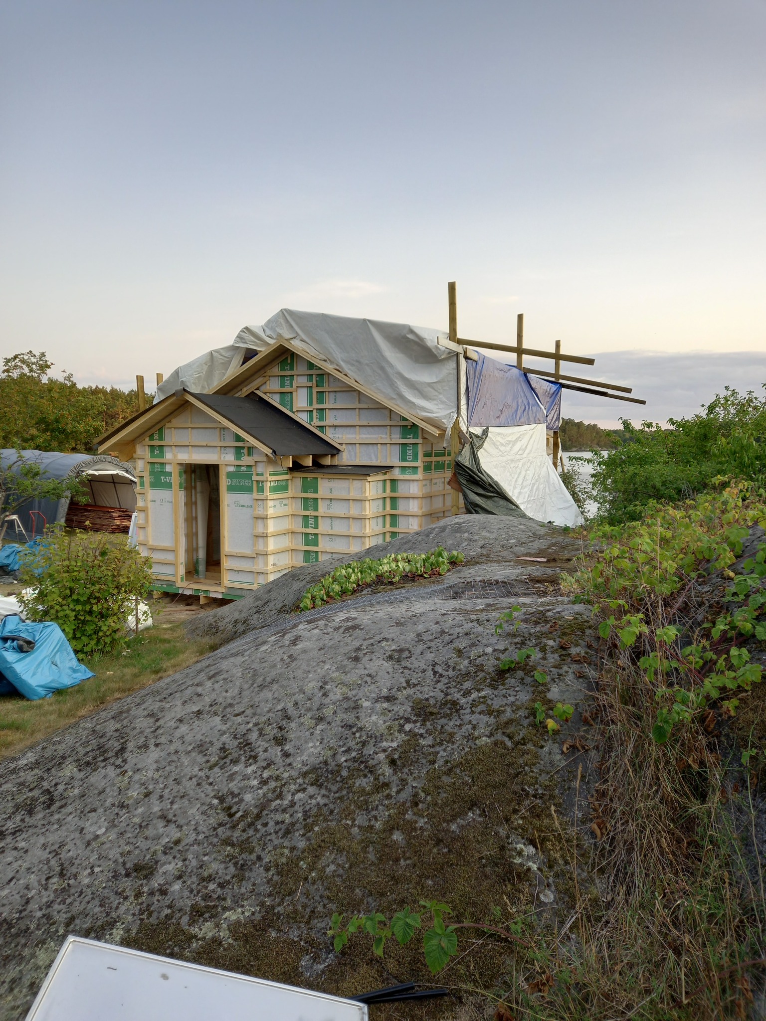 Budowa szkieletowej konstrukcji domu z widoczną izolacją, częściowo przykrytej plandeką, na nierównym terenie z dużym kamieniem.