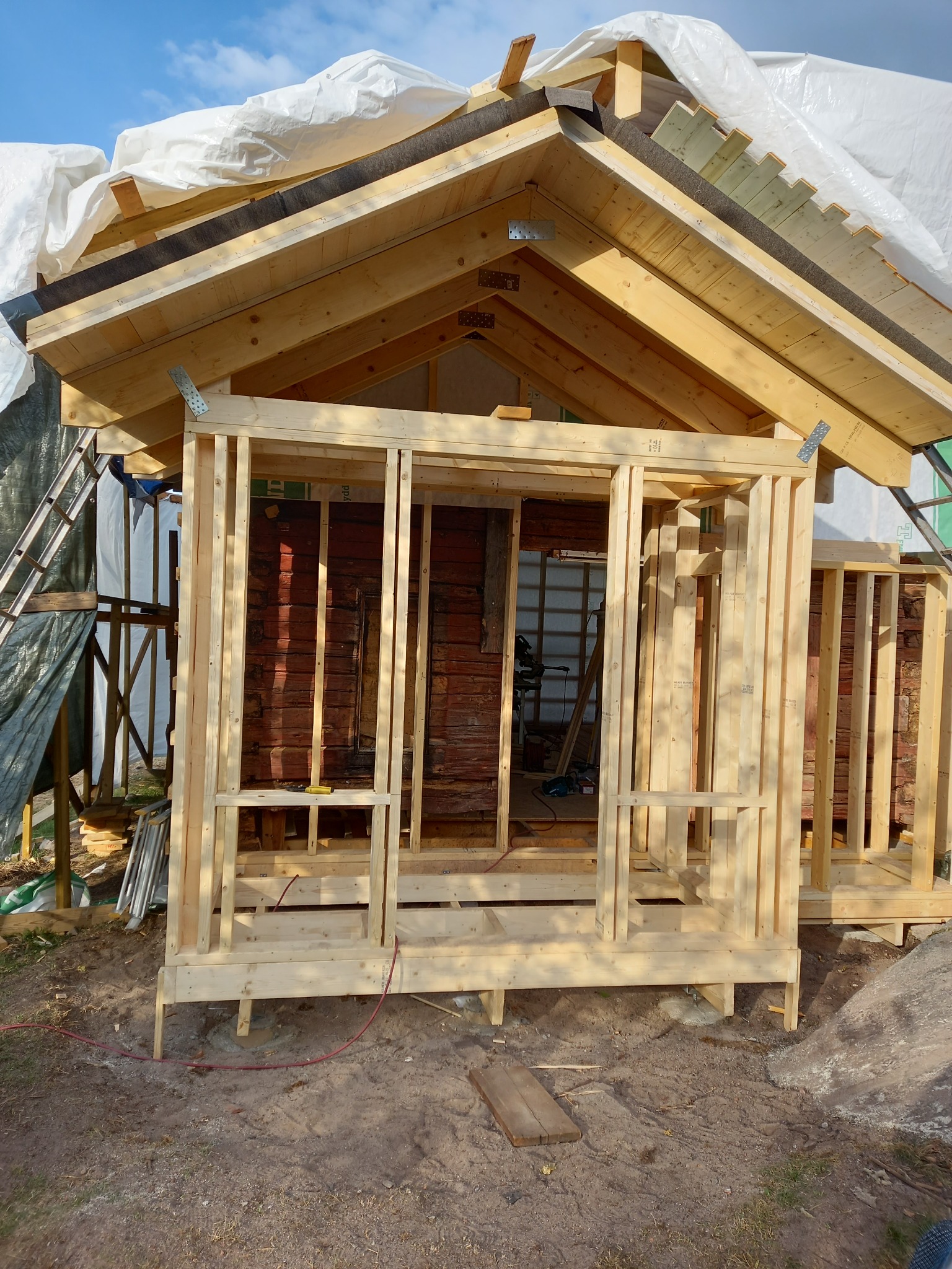 Konstrukcja szkieletowa drewnianej dobudówki z widocznym fragmentem starego budynku, zabezpieczona folią na dachu, z widocznymi elementami mocującymi z metalu.