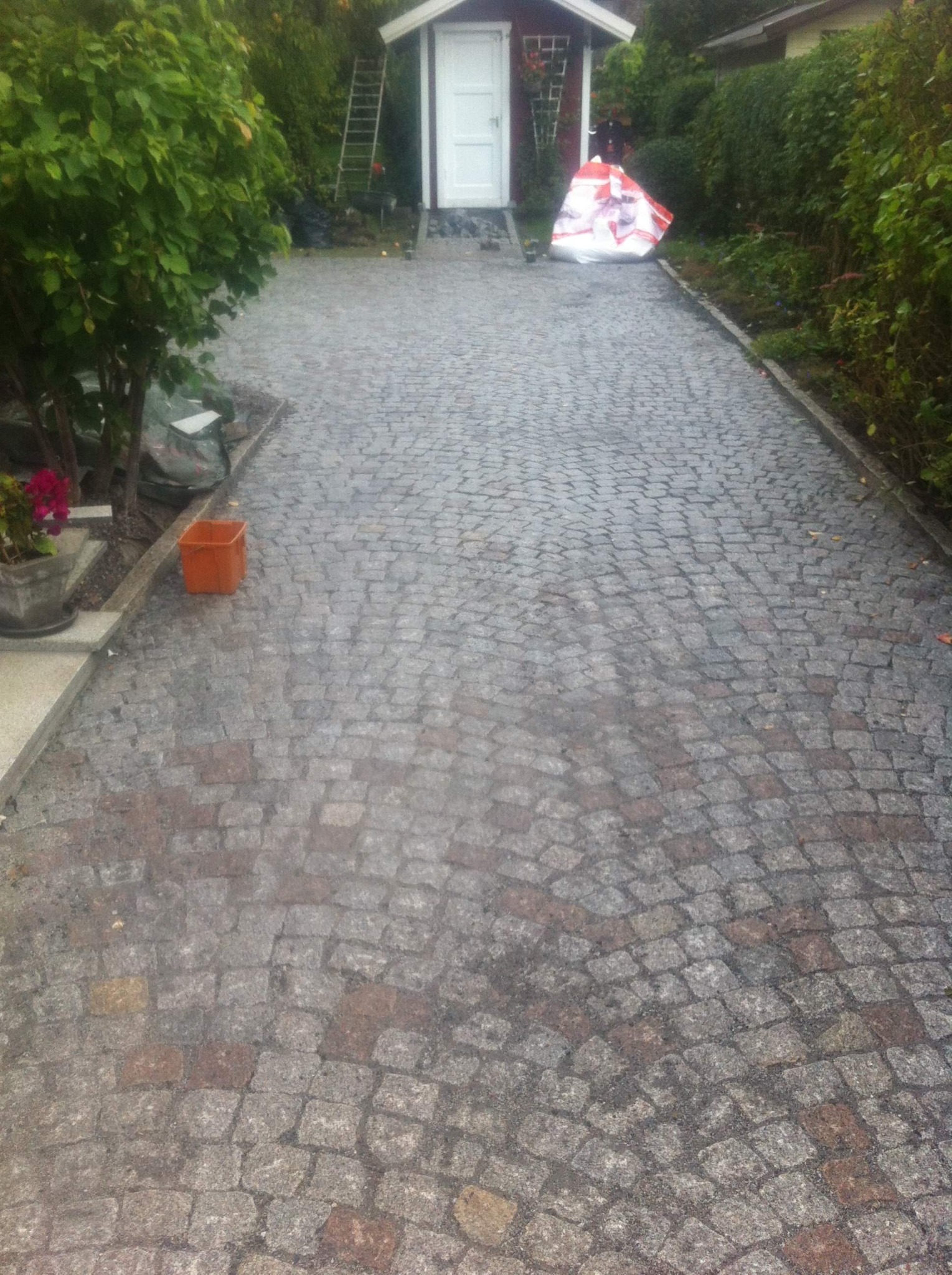 Ułożona kostka granitowa w odcieniach szarości i brązu tworzy ścieżkę prowadzącą do małego domku z białymi drzwiami, widoczny pomarańczowy pojemnik i roślinność po bokach.