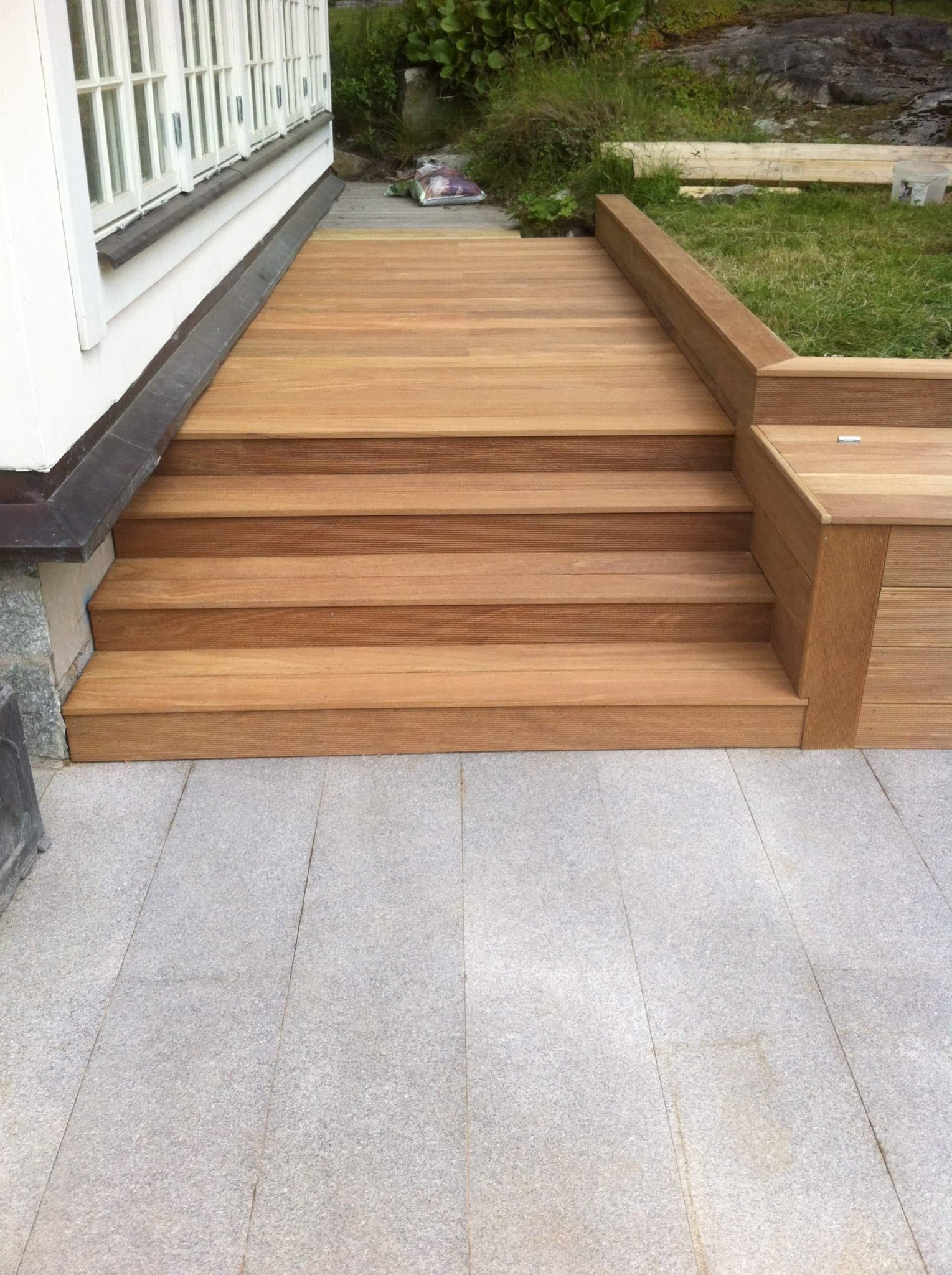 Nowoczesny taras z drewnianymi schodami i siedziskiem, wykończony szarą kostką granitową, przylegający do białego budynku z oknami.