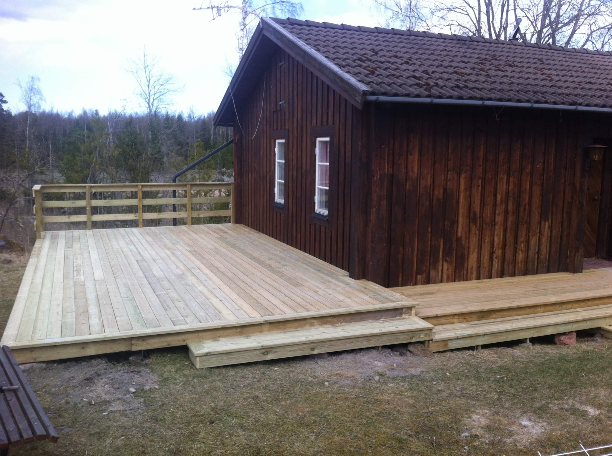 Świeżo wybudowany drewniany taras przylegający do brązowego, drewnianego domu z ciemną dachówką, widok na las w tle.