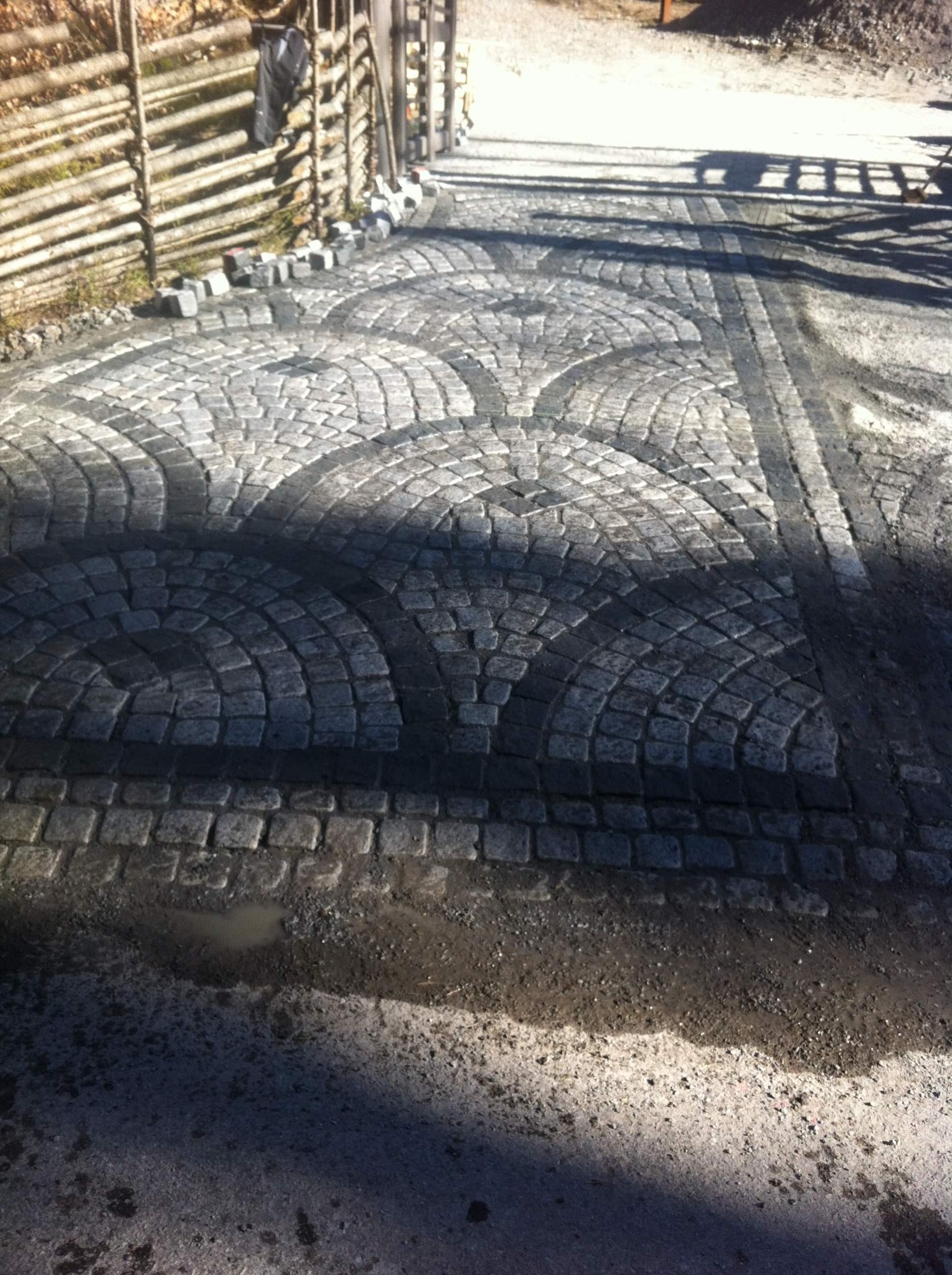 Ułożona kostka granitowa w łuki, z widocznymi śladami po deszczu i drewnianym płotem w tle.