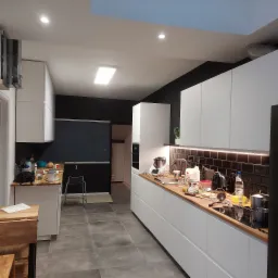 Nowoczesna kuchnia z białymi szafkami, drewnianym blatem, czarnymi płytkami na ścianie i czarną ścianą w tle, oświetlona światłem LED pod szafkami i lampami sufitowymi, w trakcie użytkowania,...