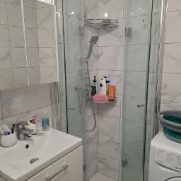 Wykończona łazienka z kabiną prysznicową ze składanymi drzwiami, umywalką z szafką i pralką, wszystko w jasnych płytkach imitujących marmur.