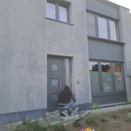 Mężczyzna w roboczym ubraniu klęczy przed wejściem do domu, malując szarą elewację. Widoczne ślady po szpachlowaniu, wiadro farby firmy Kreisel, drabina aluminiowa z lewej strony i kwiaty...