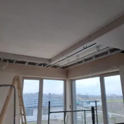 Wnętrze w trakcie montażu sufitu podwieszanego z płyt kartonowo-gipsowych, widoczne metalowe profile konstrukcyjne i częściowo zamontowane płyty, drabina oparta o ścianę, duże okna z widokiem...