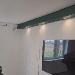 Pomalowane wnętrze: białe ściany z ciemnozielonym akcentem w górnej części, wbudowane oświetlenie punktowe, widoczny fragment sofy i telewizor z odbijającym się oknem.