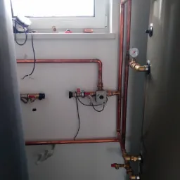 Miedziane rury instalacji grzewczej z zaworami i manometrem, zamontowane na ścianie obok zbiornika w pomieszczeniu z oknem.