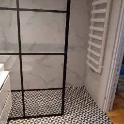 Nowoczesna łazienka z kabiną prysznicową typu walk-in z czarnymi szprosami, geometryczną czarno-białą podłogą, marmurową okładziną ścienną i białym grzejnikiem drabinkowym.