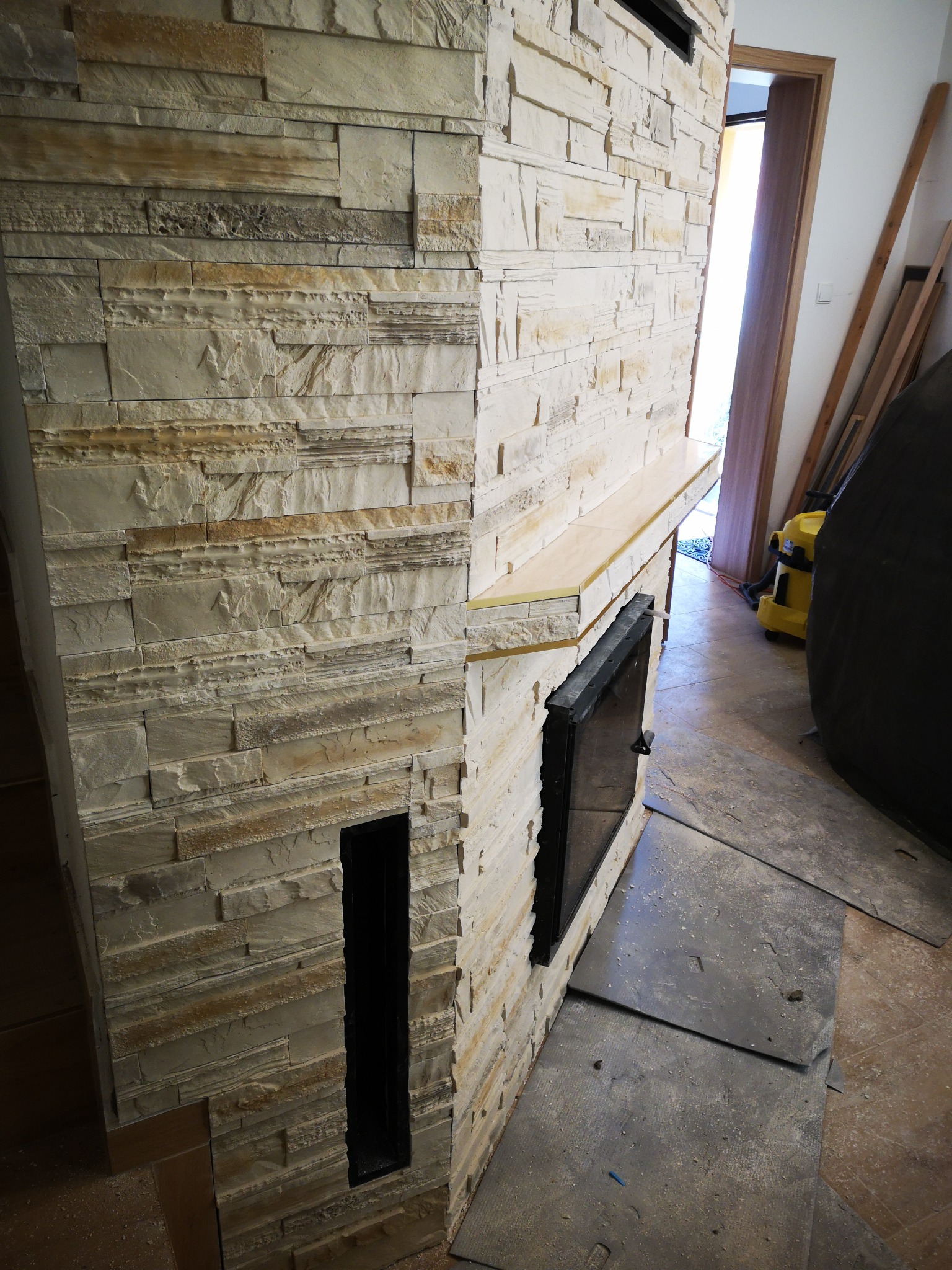 Obudowa kominka wykończona dekoracyjną, warstwową okładziną kamienną w odcieniach beżu i brązu, z widocznym wkładem kominkowym i fragmentem półki, na podłodze płyty tymczasowo zabezpieczające...