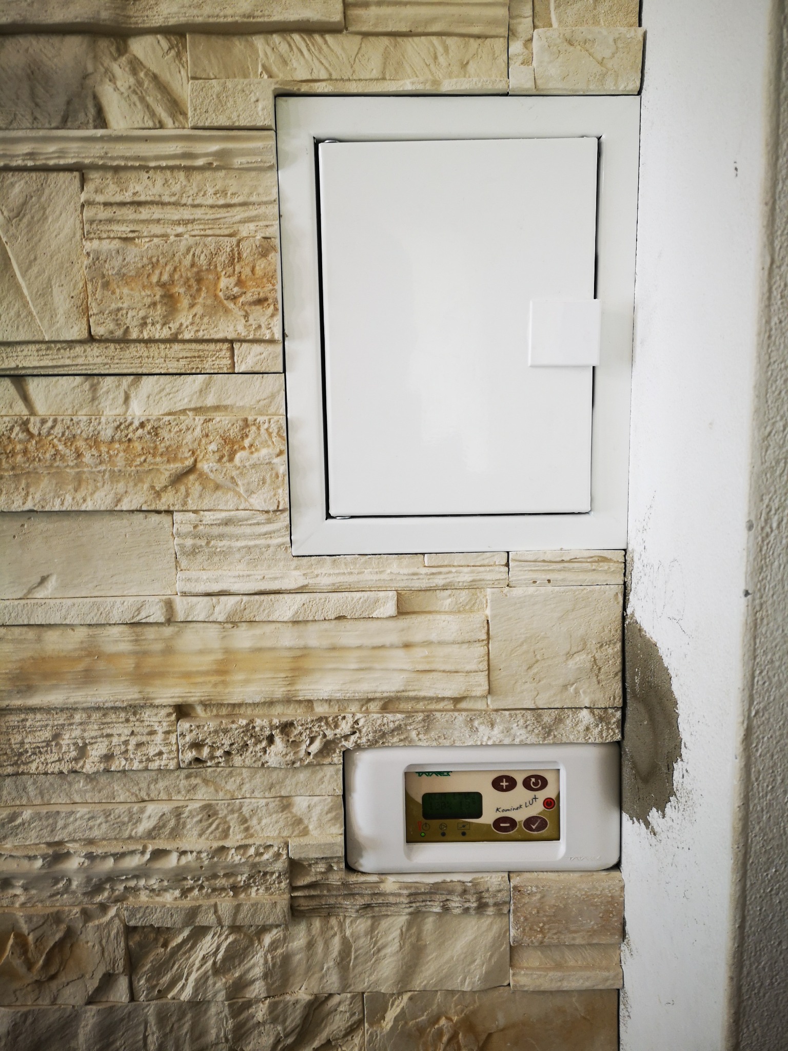 Ściana wyłożona dekoracyjnym kamieniem z wnęką na białą szafkę rewizyjną i panelem sterowania kotłem grzewczym, widoczne niedociągnięcia w wykończeniu ściany obok.