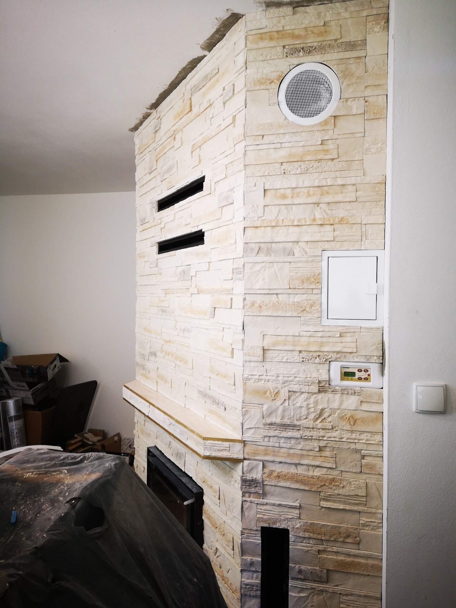 Ściana wykończona dekoracyjnym kamieniem w odcieniach beżu i bieli, z zamontowanym panelem sterowania i kratką wentylacyjną, widoczne elementy kominka przykryte folią ochronną.