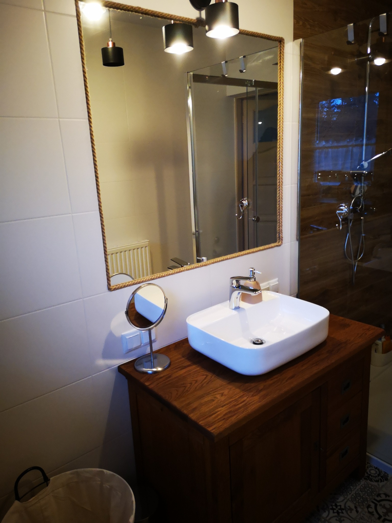 Nowoczesna łazienka z białą umywalką nablatową na drewnianej szafce, lustro w ramie z grubego sznura, kabina prysznicowa z brązowymi płytkami w tle.