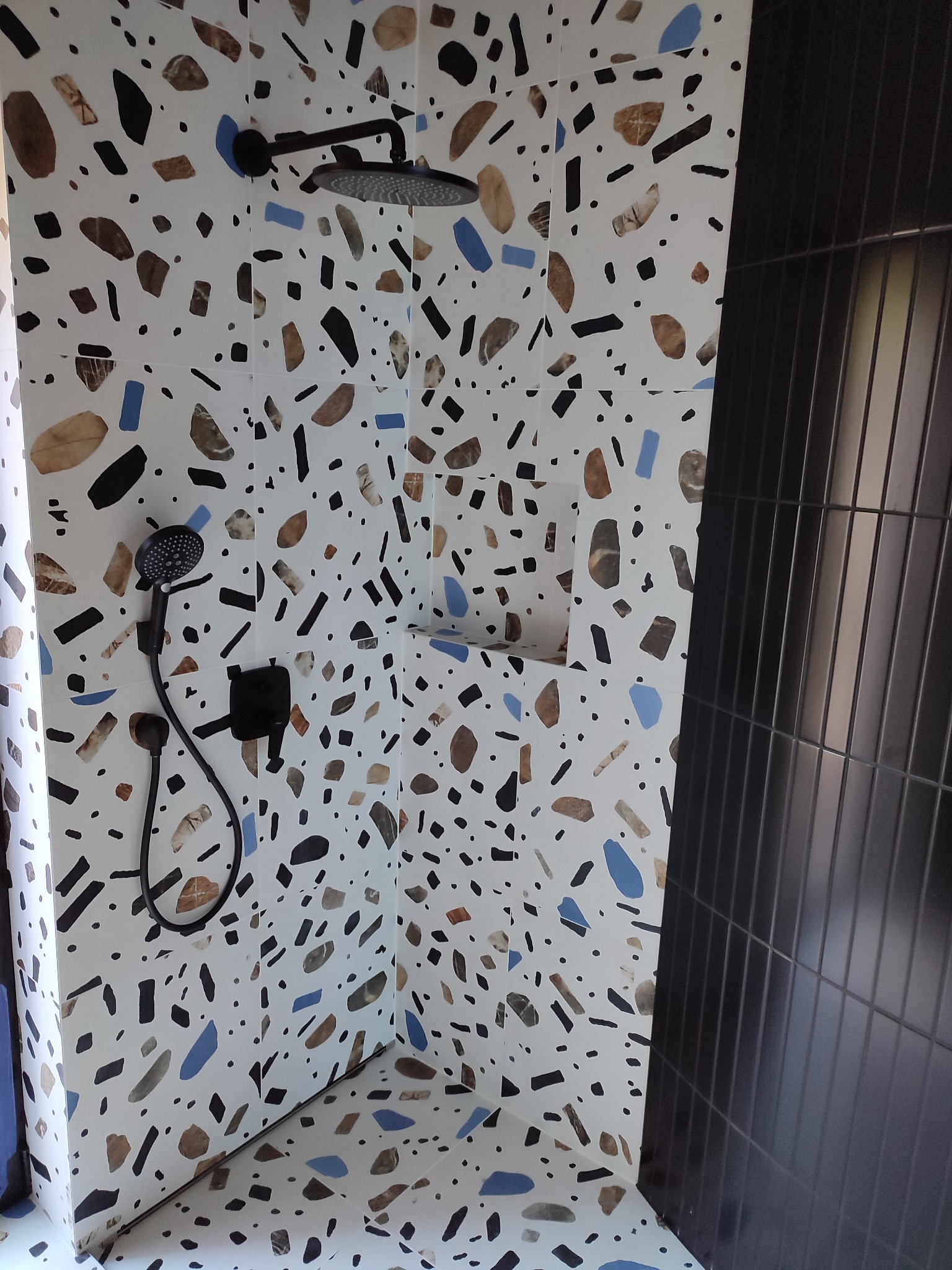 Kabina prysznicowa z oryginalną, białą okładziną w stylu lastryko z czarnymi, brązowymi i niebieskimi elementami oraz czarną armaturą i wnęką na kosmetyki.