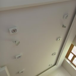 Artur kułak wykończenia wnętrz - Biały sufit z zamontowanymi, kwadratowymi lampami punktowymi w Radzyniu Podlaskim, widok z dołu, obok okno z drewnianą ramą.