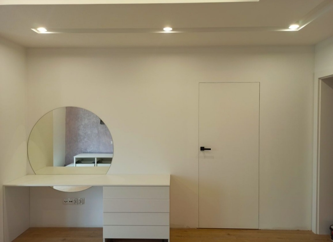 Minimalistyczna biała toaletka z okrągłym lustrem i trzema szufladami, wbudowana w ścianę, obok ukryte drzwi z czarną klamką, oświetlenie punktowe w suficie podwieszanym.