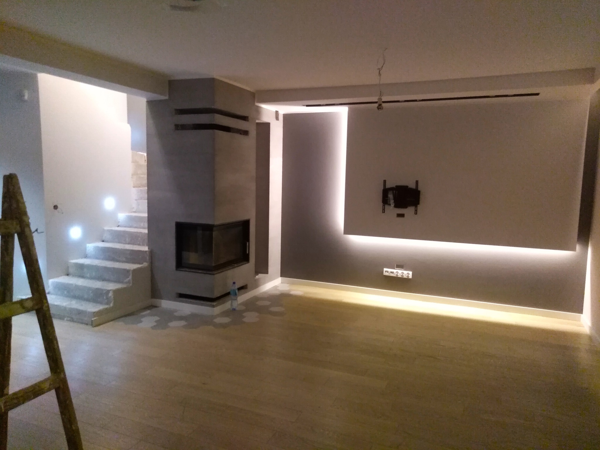 Wnętrze pokoju z kominkiem i podświetlanymi schodami w trakcie wykańczania, widoczna drabina malarska i ślady po łatach na podłodze.