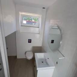 Jasna łazienka z białymi płytkami o delikatnej fakturze, wiszącą toaletą, umywalką z szafką i okrągłym lustrem z podświetleniem, widoczne niedokończone podłączenia elektryczne.