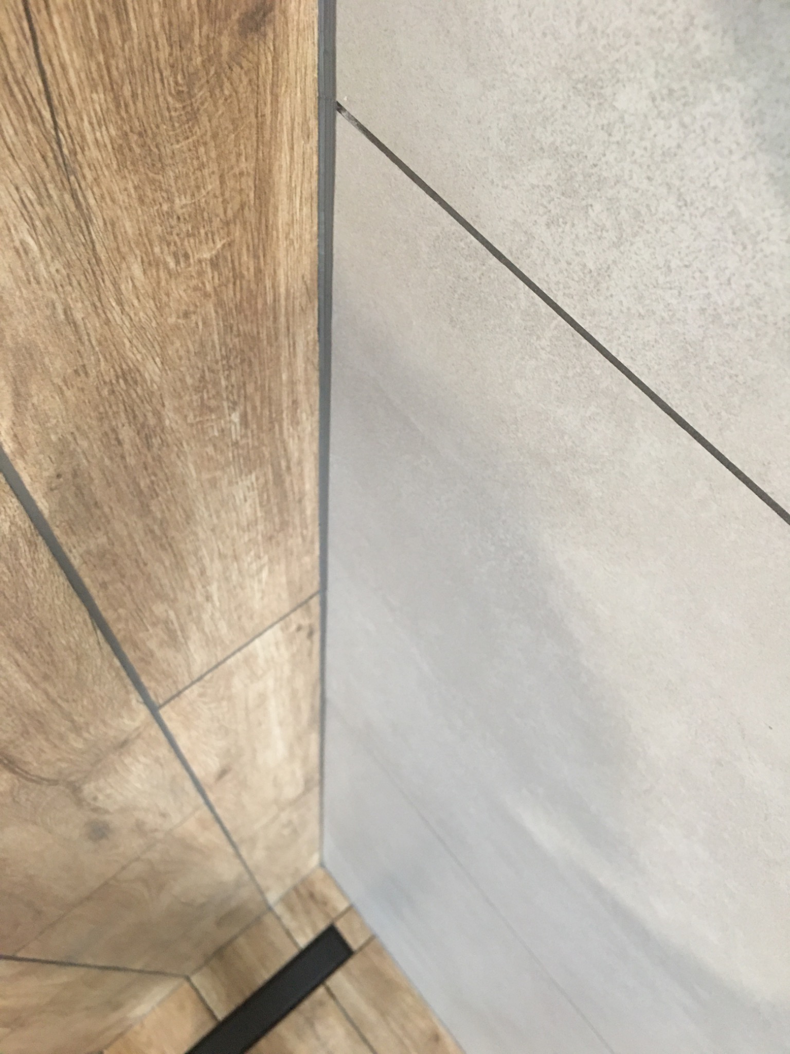 Połączenie płytek imitujących drewno na podłodze z szarymi płytkami ściennymi, widoczna czarna listwa odpływowa w rogu, fuga w kolorze szarym.