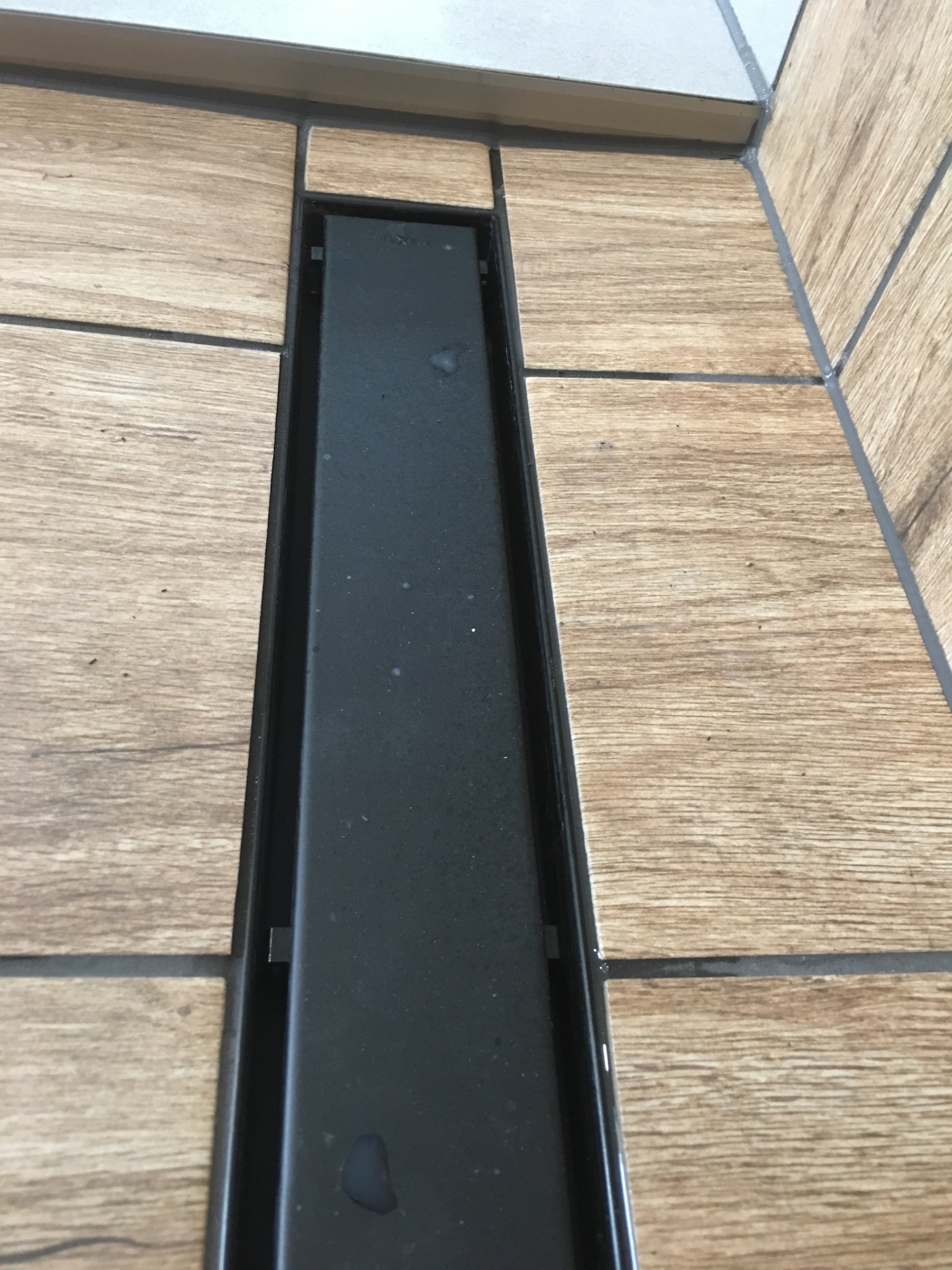 Zamontowany liniowy odpływ prysznicowy w posadzce wyłożonej płytkami imitującymi drewno, widoczne fugi i fragment ściany z jasnymi płytkami.