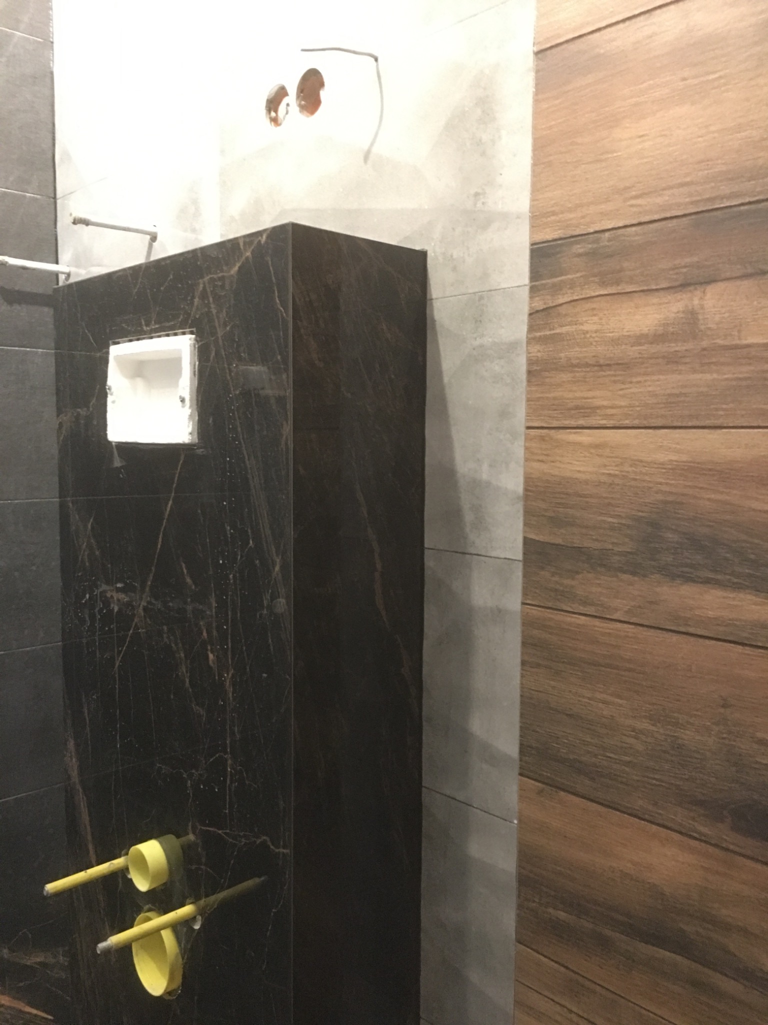 Montaż stelaża podtynkowego WC z czarną płytą imitującą marmur, widoczne rury odpływowe w kolorze żółtym oraz fragment ściany wykończonej panelami drewnopodobnymi.