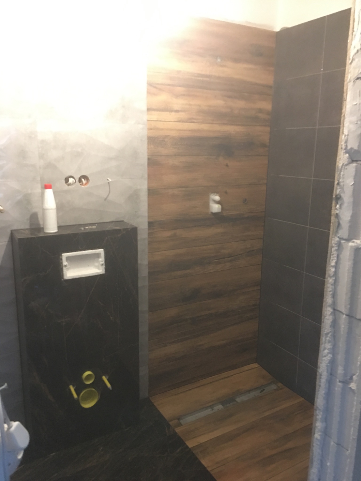 Częściowo wykończona łazienka z panelami ściennymi imitującymi drewno, szarą ścianą z dekoracyjnym wzorem, podtynkową toaletą z czarnym marmurkowym wykończeniem i widocznymi elementami instalacji.