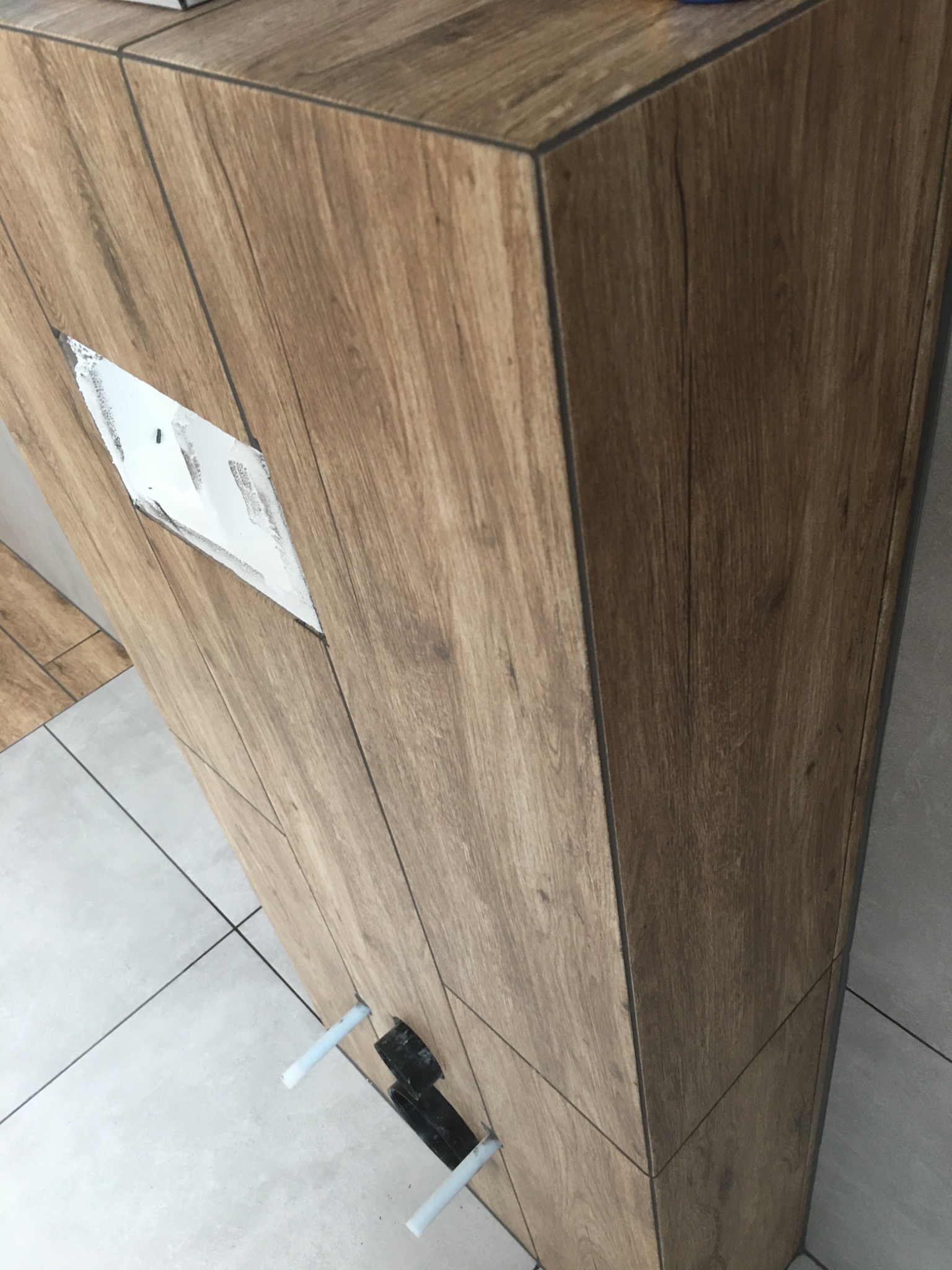 Obudowa stelaża WC z płytek imitujących drewno, częściowo wykończona, z widocznym otworem na przycisk spłukujący i rurami doprowadzającymi wodę, na tle szarej podłogi z płytek.