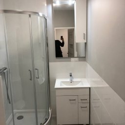 Usługi remontowo wykończeniowe - Nowoczesna łazienka z kabiną prysznicową, białą umywalką z szafką i lustrem z oświetleniem, widoczne odbicie osoby robiącej zdjęcie telefonem.