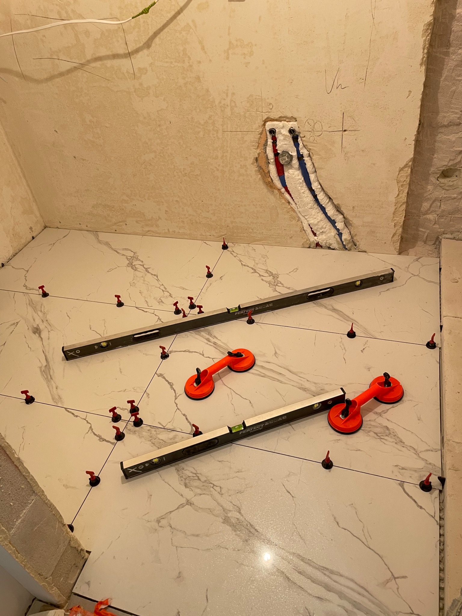 Układanie białych płytek imitujących marmur na podłodze łazienki z użyciem poziomnic i przyssawek do płytek. Widoczne kliny i system poziomowania płytek.