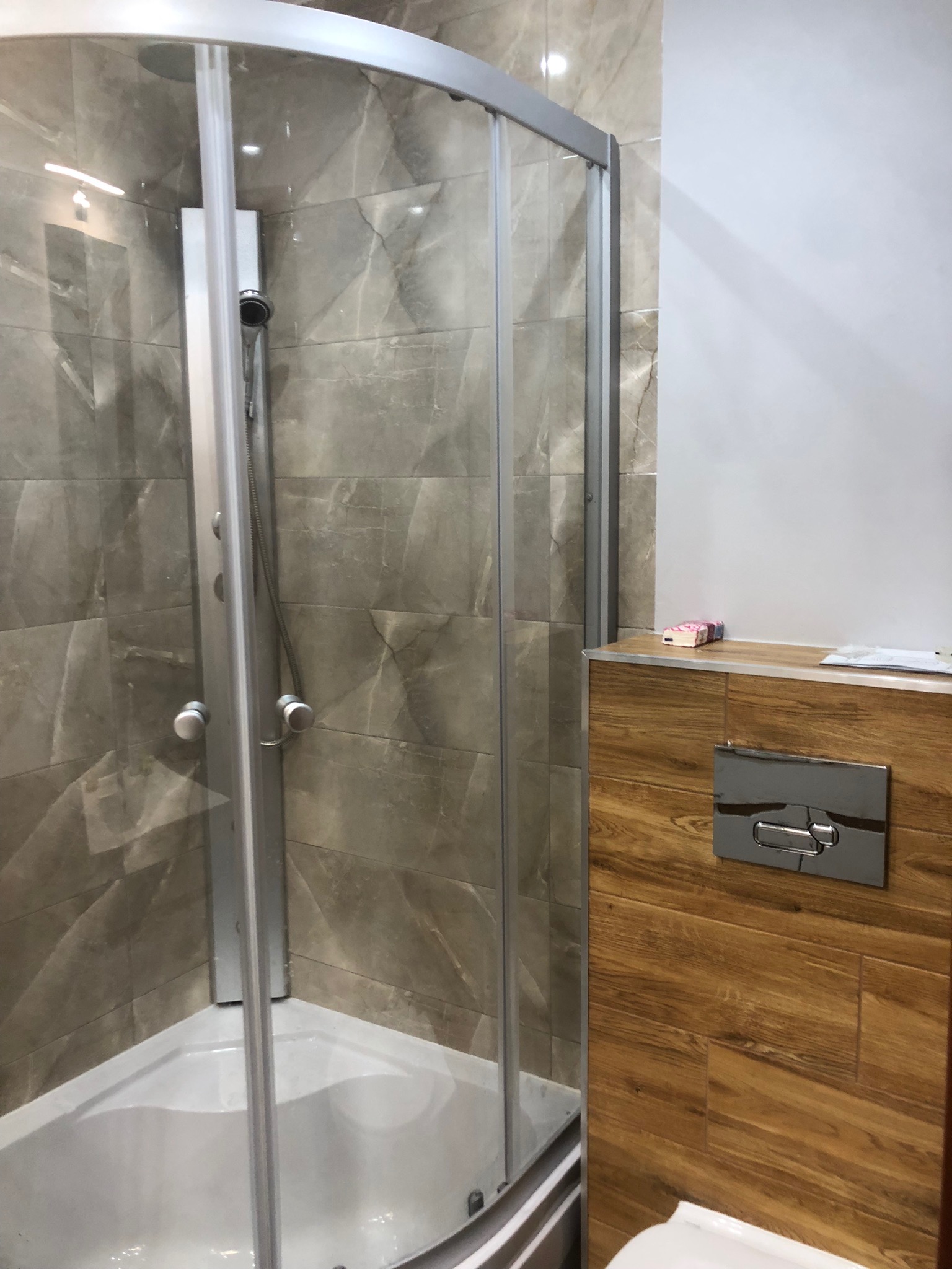 Narożna kabina prysznicowa z szarymi płytkami imitującymi marmur i drewnopodobną zabudową geberitu