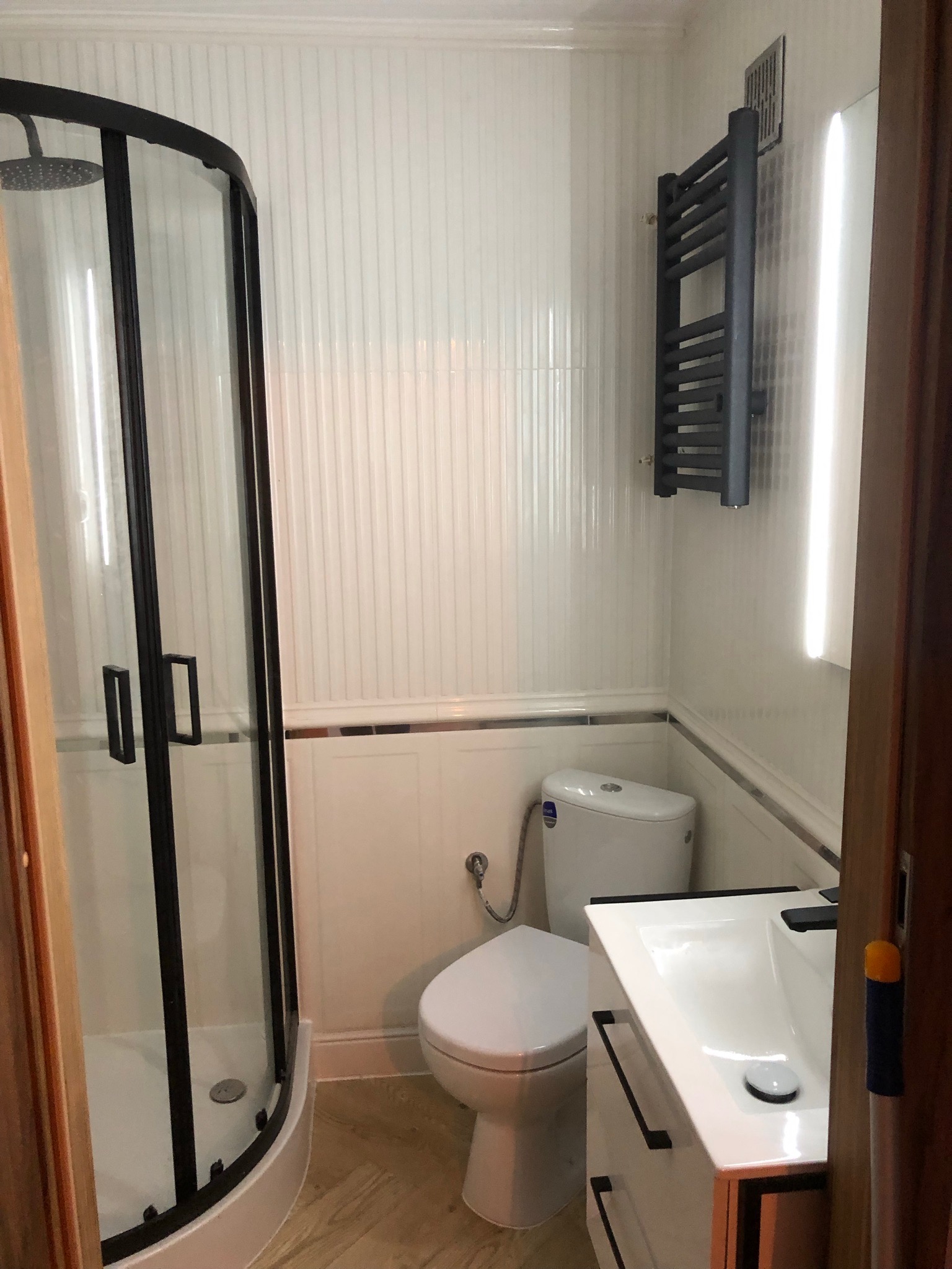 Nowoczesna łazienka z kabiną prysznicową z czarnymi ramami, wiszącym grzejnikiem drabinkowym w kolorze grafitowym, białą toaletą i umywalką z szafką.