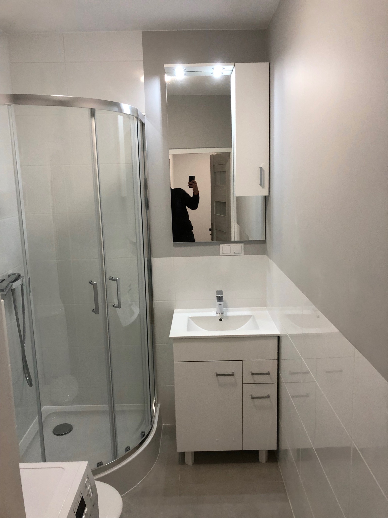 Nowoczesna łazienka z kabiną prysznicową, białą umywalką z szafką i lustrem z oświetleniem, widoczne odbicie osoby robiącej zdjęcie telefonem.