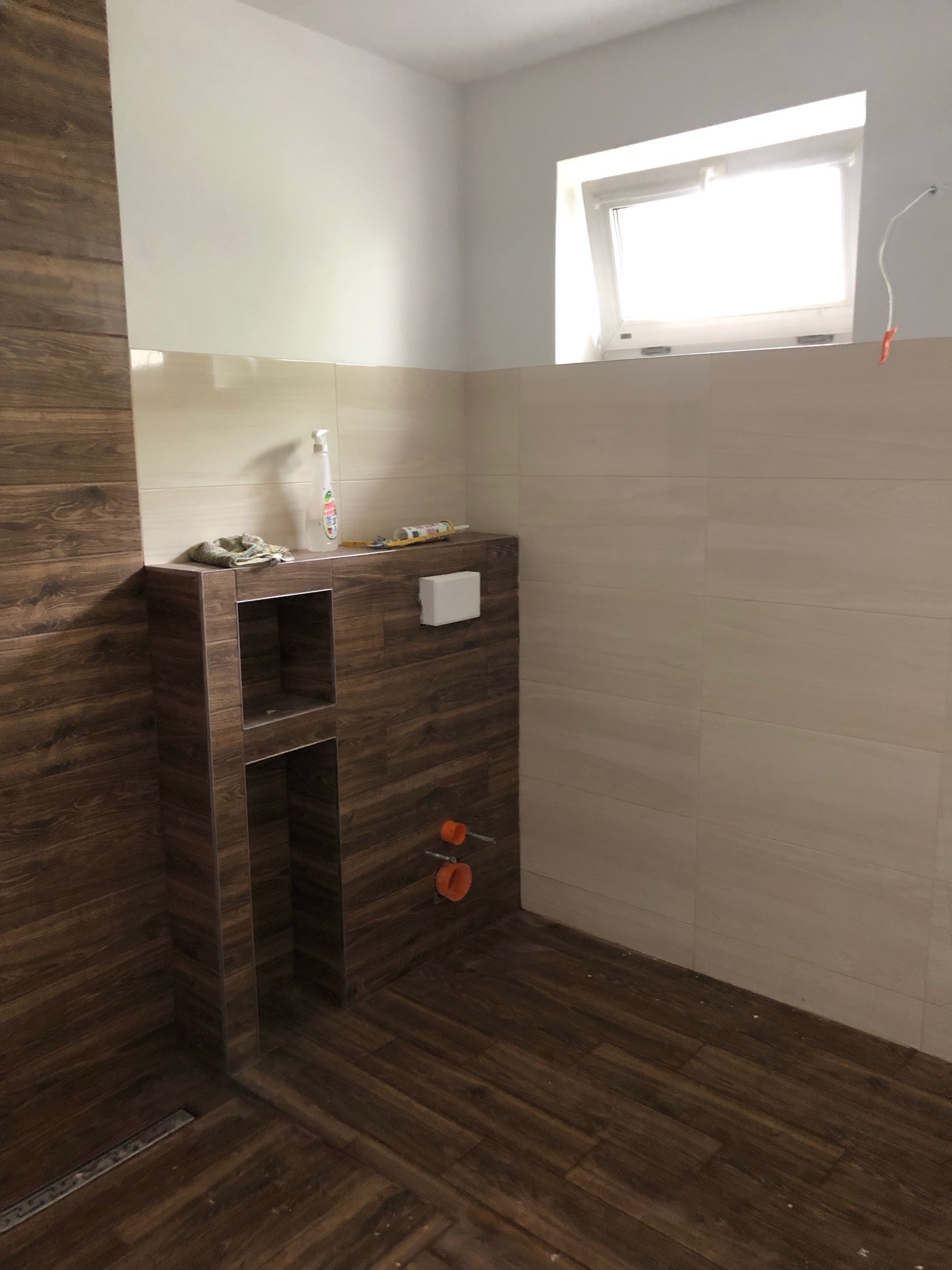 Wykończona łazienka z drewnopodobnymi płytkami na podłodze i ścianach, zabudowana toaleta z wnękami, widoczne rury instalacji, okno w górnej części ściany.