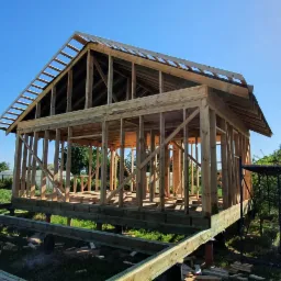 Surowa konstrukcja drewniana domu szkieletowego na podwyższeniu, widoczna na tle błękitnego nieba i zieleni ogrodu, z fragmentem szklarni po lewej stronie.