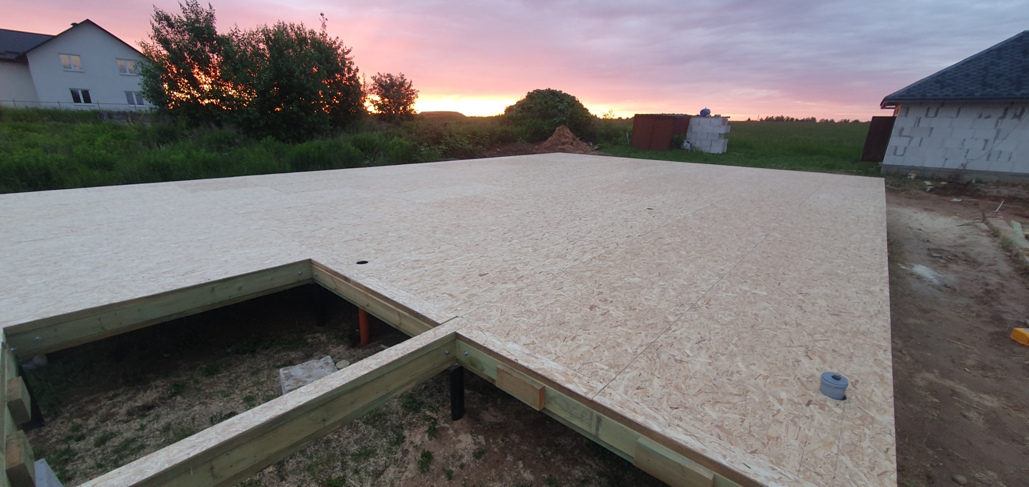 Fundament domu z płyty OSB na drewnianej konstrukcji szkieletowej, widok z góry, w tle budynek w trakcie budowy i zachodzące słońce.